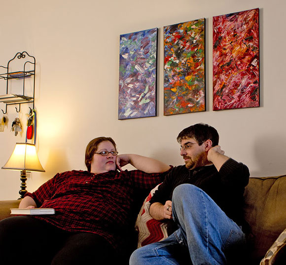 Havens sits on the couch with his girlfriend. Colorful paintings hang on the wall above the couch.